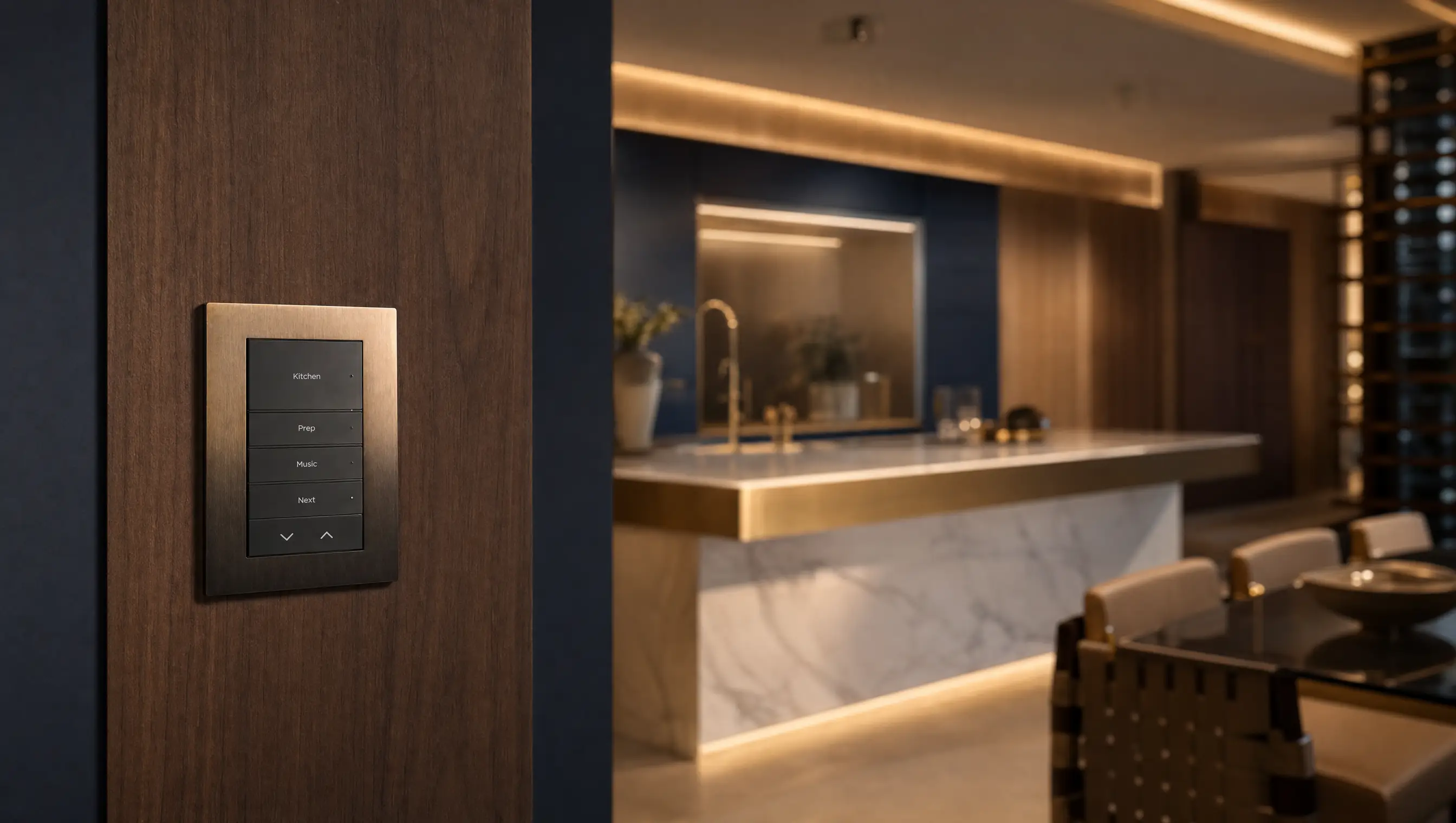 A bronze engraved Control4 keypad on a walnut panel beside a marble and brass kitchen at dusk
